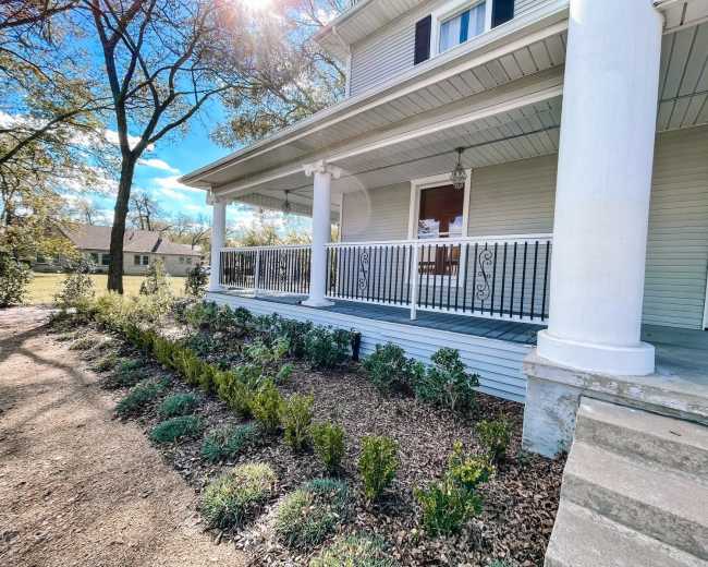 The image shows a porch with white columns and railings, flanked by neatly arranged shrubs and mulch, in a residential setting under a blue sky.