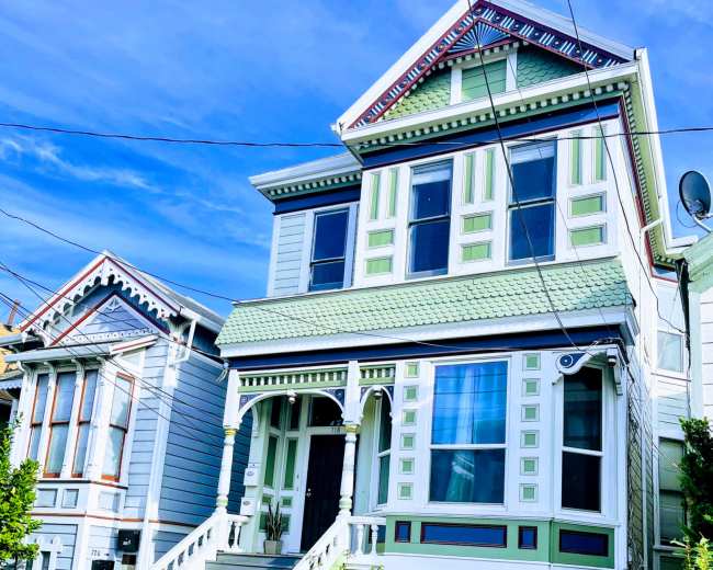 A Victorian-style house with green and white trim, featuring a front porch and decorative details, stands on a residential street.