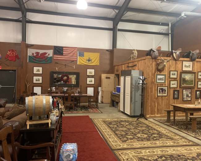 The scene depicts a spacious room with wooden walls, a dining area featuring a long table and chairs, several framed artworks, and decorative flags hanging above, along with seating arrangements and a faux rustic barrel.