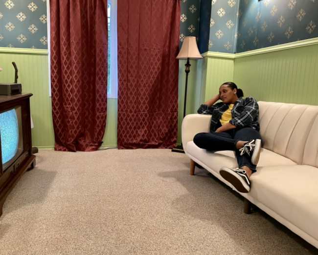 A person sits on a white couch in a room with green and blue walls, watching an old television while a lamp stands nearby.