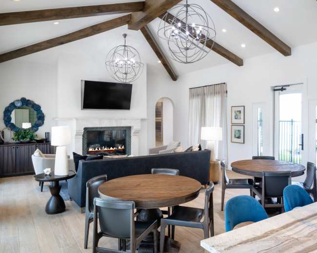 The image shows a modern living space with high beamed ceilings, a fireplace, multiple dining tables, and large windows that provide natural light.