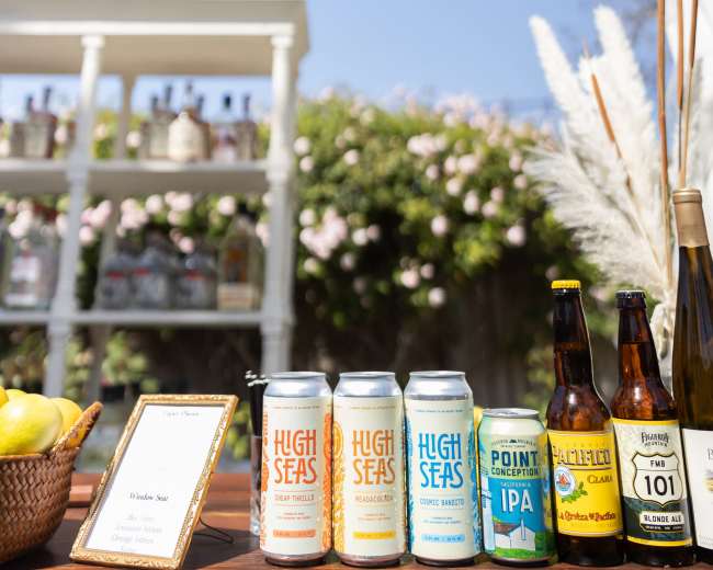 A selection of craft beers is displayed on a table under a clear blue sky, with a background of a shelf holding various bottles.