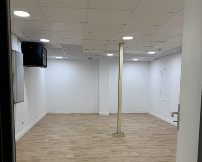 The image shows a spacious, empty room with wooden flooring, a central support pillar, and ceiling lights.
