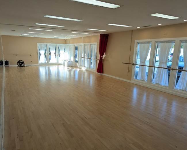 The image shows a spacious, well-lit dance studio with wooden flooring, large mirrors, and white curtains.