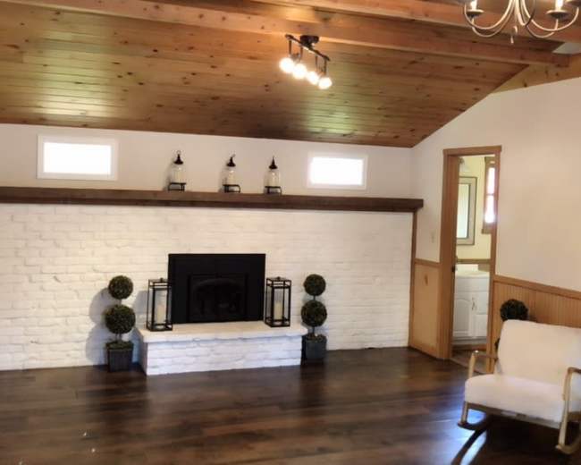 A spacious living room features a white brick fireplace, wooden ceiling beams, and light hardwood flooring.