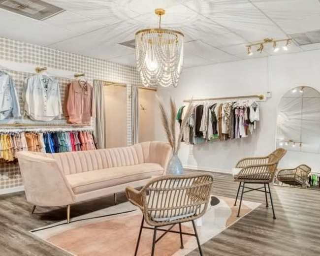 The image features a stylish boutique interior with a pink velvet sofa, two woven chairs, a large mirror, and racks of clothing displayed on the walls.