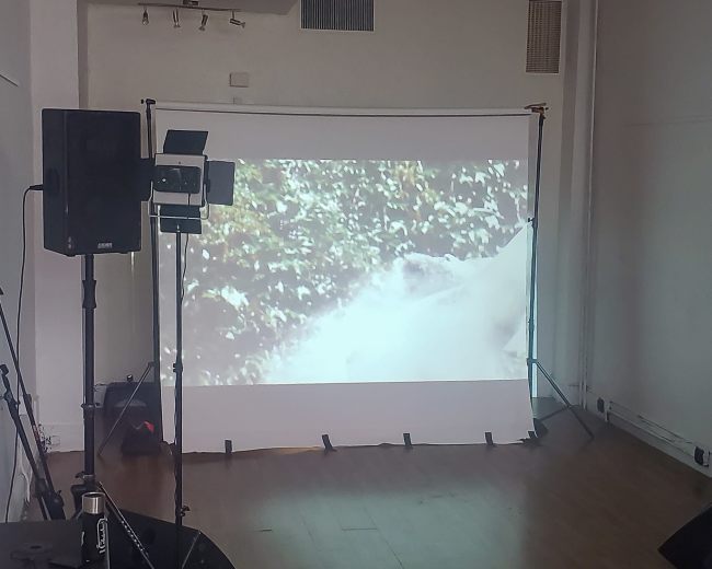 A projection screen displaying a video is set up in a sparsely furnished room, with sound equipment and a bucket on the floor.