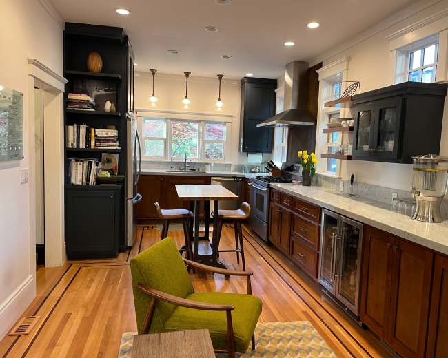 A modern kitchen features dark wood cabinets, a marble countertop, and a small dining table, with ample natural light from windows.