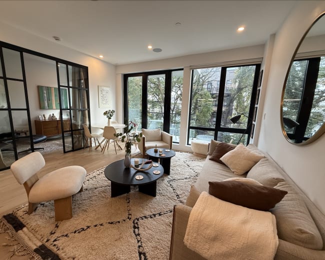 The image shows a modern living room with a light-colored sofa, a round coffee table, and large windows that offer a view of greenery outside.