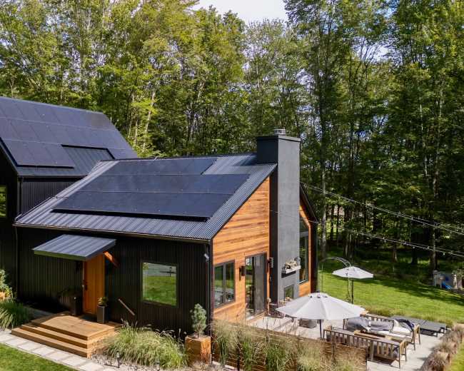 The image shows a modern black and wood house with solar panels on the roof, situated in a wooded area with a patio and outdoor furniture.
