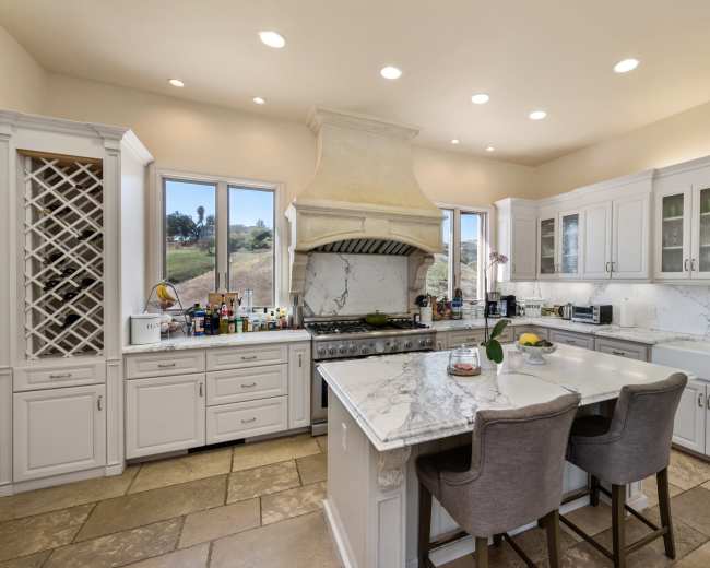 A spacious kitchen features white cabinetry, a marble countertop island, and large windows overlooking a scenic landscape.