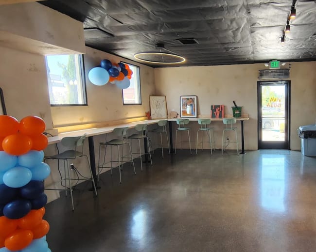 The image shows a modern interior of a café or event space featuring a bar area with seating, decorated with orange and blue balloons at both ends.