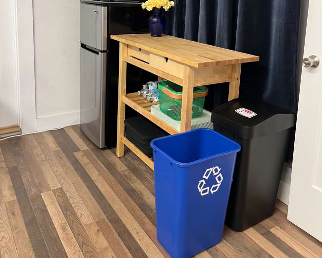 The image shows a small kitchen area featuring a microwave, a mini-refrigerator, a wooden table with flowers, and two trash bins, one for recycling.