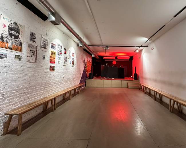 The image shows a narrow performance space with wooden benches on either side and a small stage lit with red lighting at the far end.
