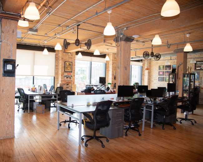 The image shows a modern office space featuring multiple desks with computers, wooden floors, exposed beams, and large windows allowing natural light.