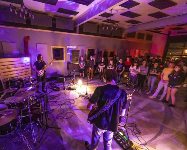 A band performs on a low-lit stage while an audience watches intently in a venue filled with purple lighting.