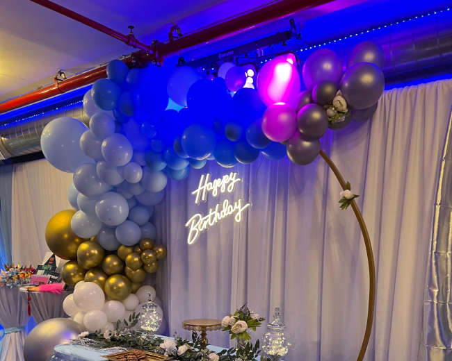 A decorative birthday setup features a table covered with a light blue ruffled cloth, adorned with a neon "Happy Birthday" sign and an arch of colorful balloons.