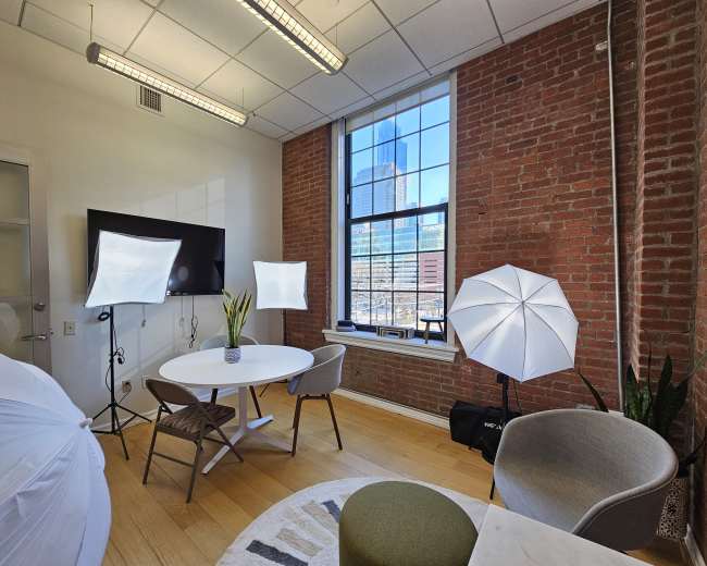 The image shows a photography studio with a round table, chairs, lighting equipment, and large windows displaying a cityscape.