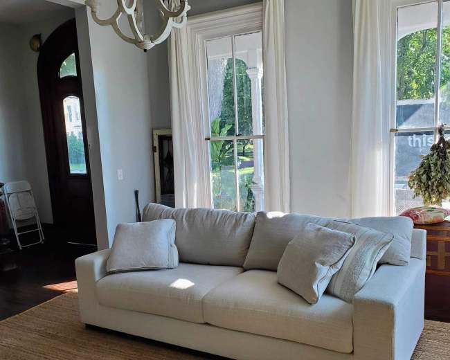The image shows a light-colored sofa with two cushions, placed on a woven rug in a room with large windows and a doorway.