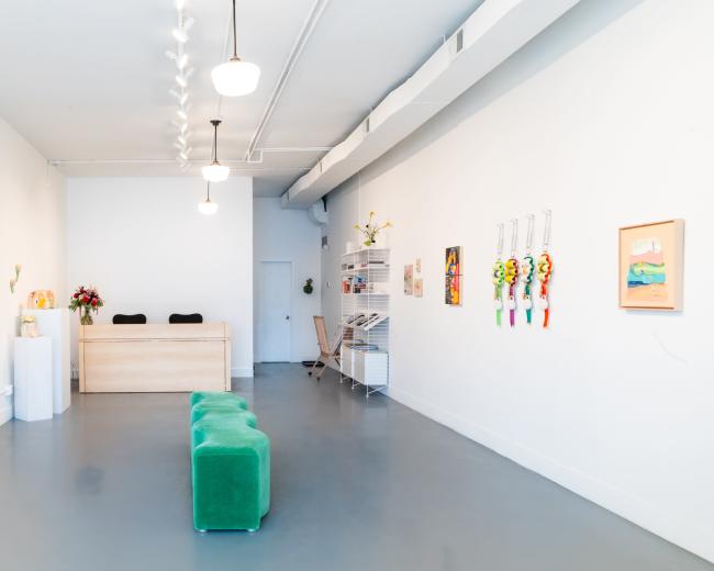 The image shows a bright, minimalist art gallery with a reception desk, colorful paintings on the walls, and a green seating area in the center.