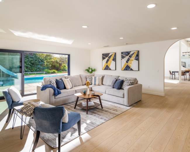 A spacious living room features a light-colored sectional sofa, two blue chairs, and large windows overlooking a pool area.