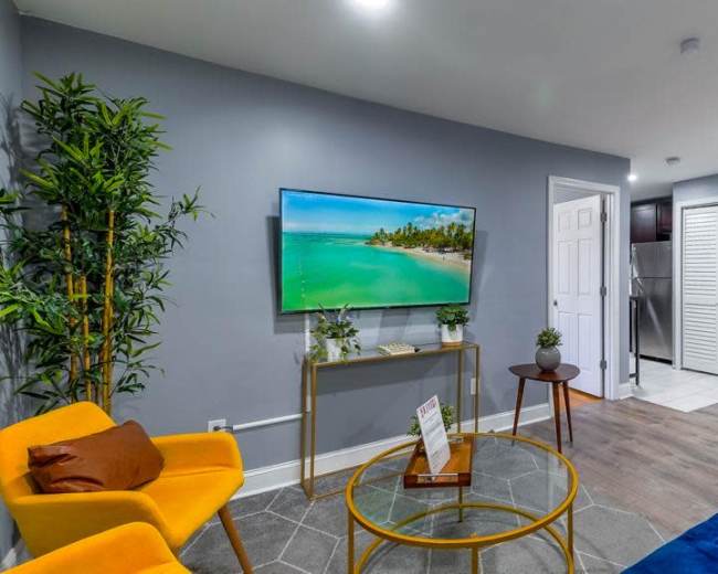 A modern living room featuring a large TV mounted on the wall, two colorful chairs (one yellow and one blue), a round coffee table, and a doorway leading to a kitchen area.
