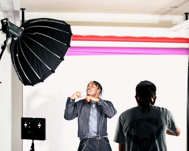 A model poses in front of a backdrop while a photographer adjusts lighting equipment in a studio.