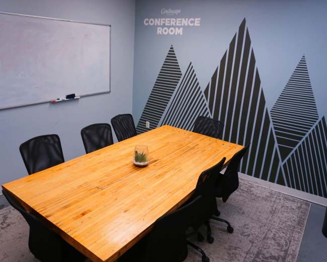 The image shows a conference room featuring a long wooden table surrounded by black office chairs, with a whiteboard on one wall and a mountain-themed mural on another.