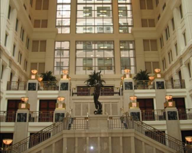 The image shows a grand interior of a building featuring a large archway, a central staircase, and decorative plant elements.