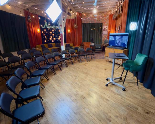 The image shows a well-decorated room set up for a presentation, featuring rows of black chairs, a computer on a mobile stand, and a backdrop adorned with colorful curtains and lights.