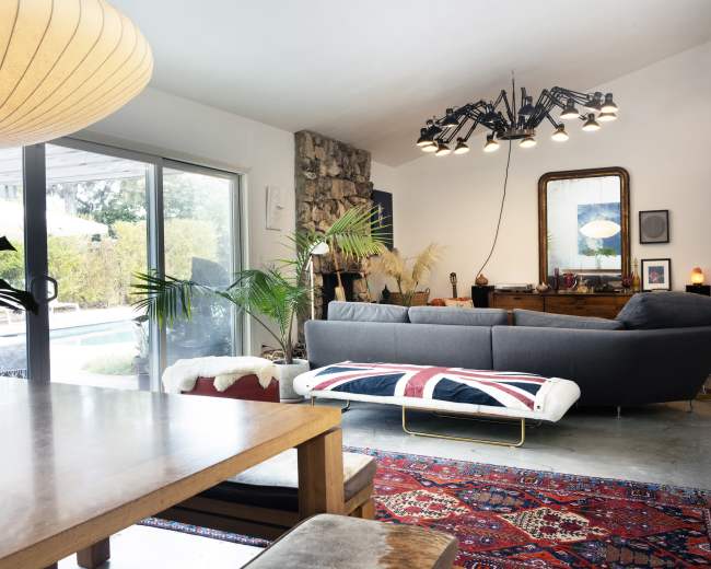 The image shows a modern living room with a gray sofa, a wooden dining table, and large glass doors leading to an outdoor pool area, decorated with plants and a colorful area rug.