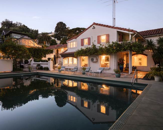 The image shows a tranquil swimming pool surrounded by lush greenery and illuminated buildings at dusk.