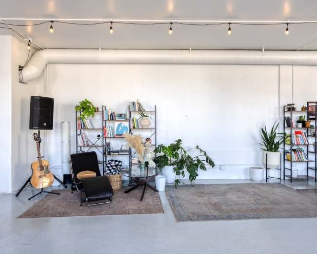 The image shows a spacious room with shelves filled with books and plants, a guitar, a chair, and decorative rugs on a light-colored floor.