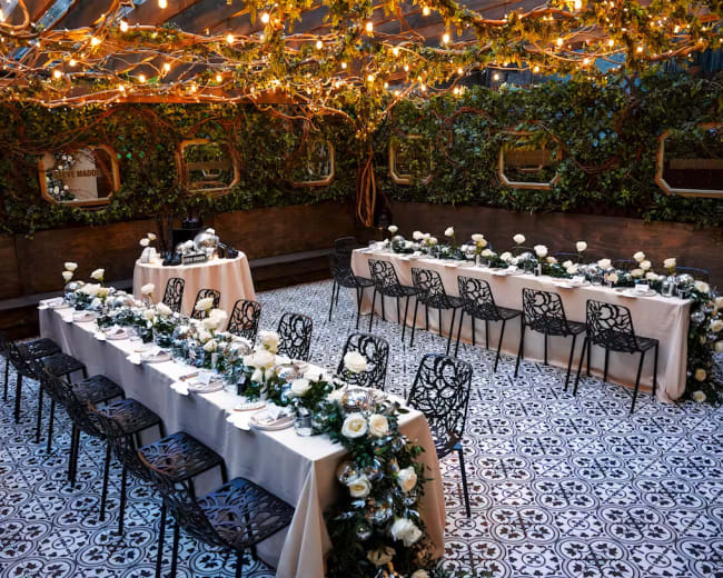 A dining area is arranged with long tables adorned with white tablecloths and floral centerpieces under warm string lights and green foliage.