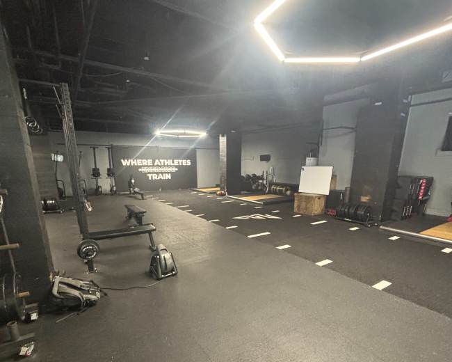 The image shows an empty gym space with black flooring, various fitness equipment, and a motivational wall that reads "WHERE ATHLETES TRAIN."