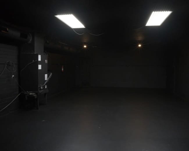 The image shows a dimly lit, empty room with black walls and a smooth black floor, featuring overhead fluorescent lights and a small machine in one corner.