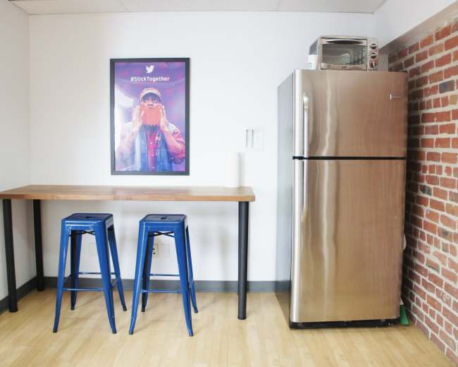 A small kitchen area features a metallic refrigerator, a microwave above it, a wooden table with two blue stools, and a framed poster on the wall.