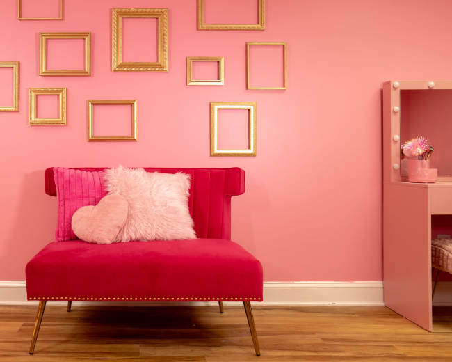 The image shows a vibrant pink room featuring a red velvet chair, adorned with a heart-shaped pillow and surrounded by empty gold-framed pictures on the wall.