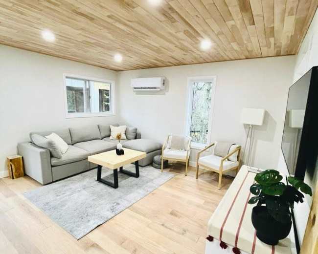 The image shows a cozy living room with a gray sofa, two chairs, a coffee table, and wood-paneled ceilings.