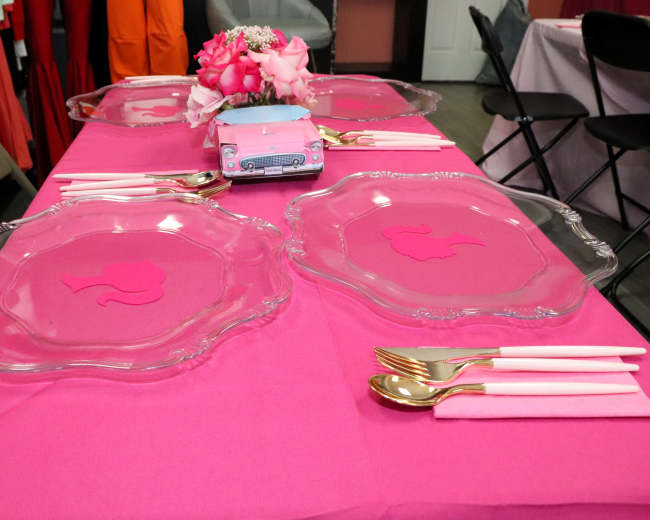 A pink table is set with glass plates and gold utensils, featuring a centerpiece of flowers and a decorative item.