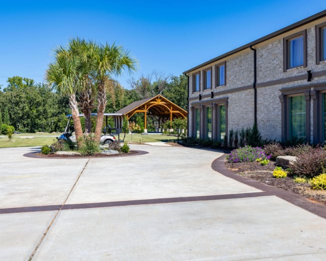The image shows a landscaped property with a building on one side, a curved driveway, and a wooden gazebo in the background.