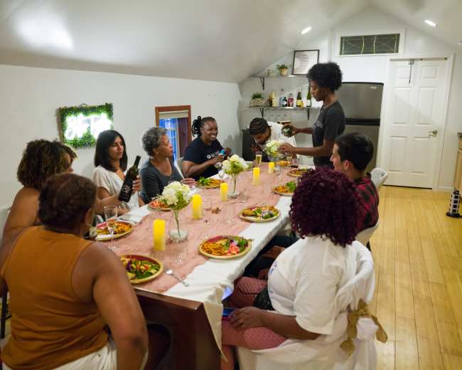 A group of people gather around a table set with food and candles in a well-lit room.