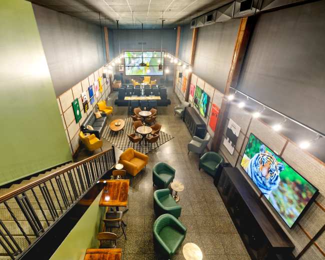 The image shows an interior space with a modern café or lounge setup, featuring a variety of seating arrangements, artworks on the walls, and large screens displaying images.