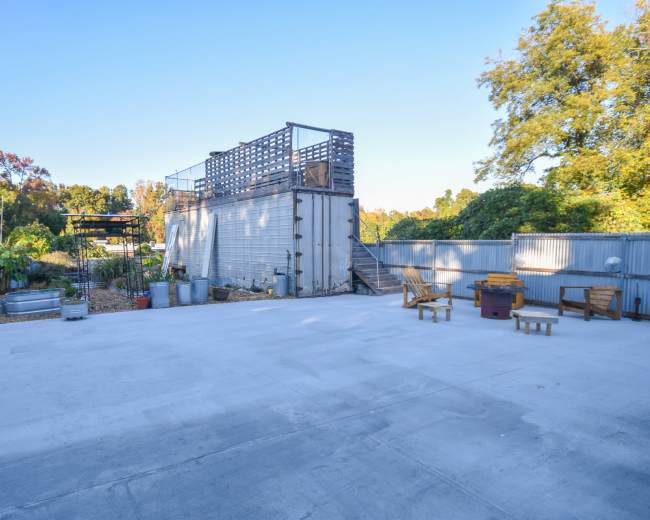 The image shows a spacious outdoor area with a concrete surface, surrounded by metal fencing, featuring a two-story structure, wooden benches, and landscaped greenery in the background.