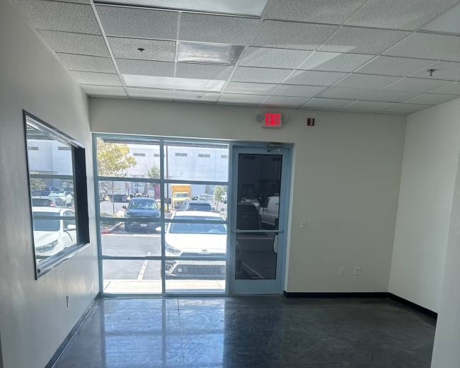 The image shows an empty office space with a large window and a glass door leading to a parking area.