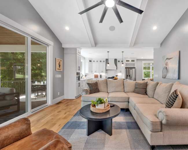 The image shows a modern living room featuring a large sectional sofa, a round coffee table, and an open layout with a view of a kitchen area in the background.