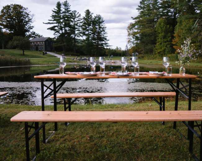 A long wooden table set for a meal is positioned beside a pond, surrounded by trees and a cloudy sky.