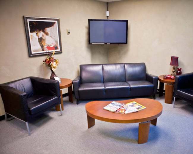 The image shows a waiting area with a black leather couch, two black armchairs, a round wooden coffee table with magazines, and a decorative lamp, all against a painted wall with a framed painting and a wall-mounted TV.