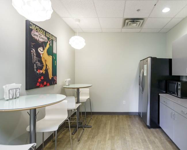 The image shows a modern break room with a round table, four high-backed stools, a refrigerator, a microwave, and a wall-mounted artwork.