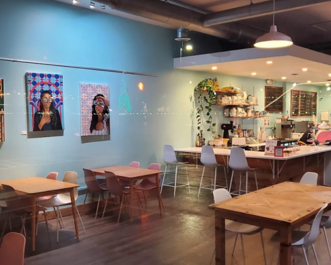 The image shows a café interior with colorful artwork on the walls, wooden tables, and a counter displaying various food and drink items.